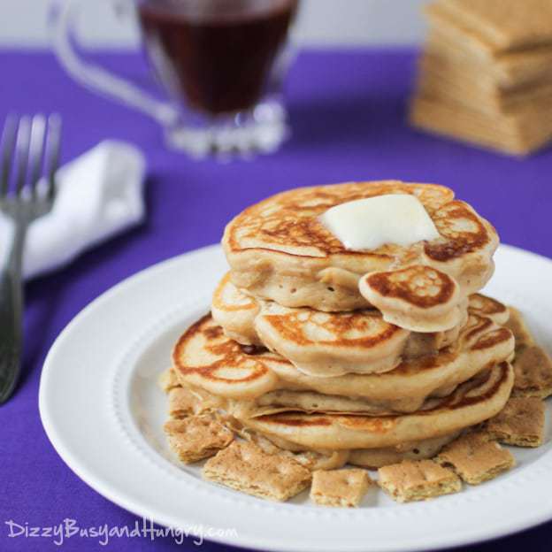 Side view of stacked graham cracker pancakes topped with a pad of butter with a cup of coffee, graham crackers, and a fork in the purple background.
