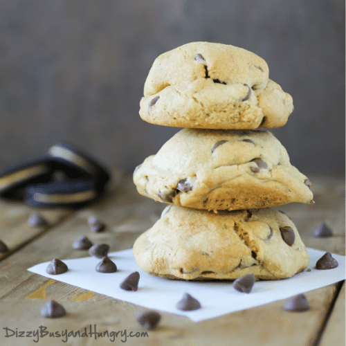 Side view of three PB Oreo chocolate chip cookies stacked on a white paper on a wooden surface with chocolate chips and Oreos in the background. 