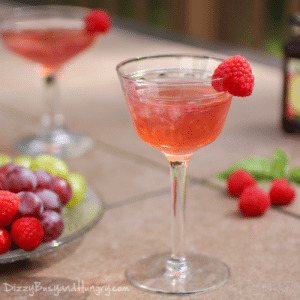 Side view of raspberry gimlet in a cocktail glass with a raspberry on the rim with grapes and raspberries on the side.