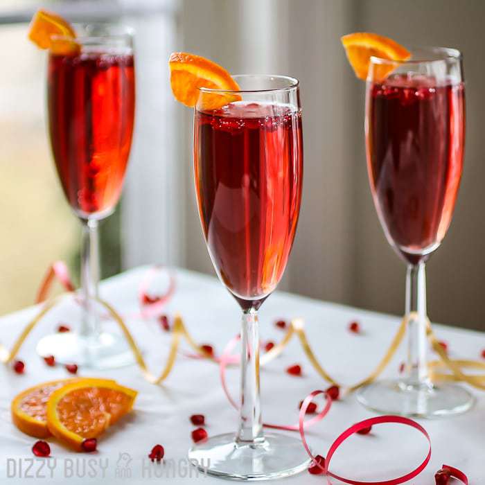 Side view of three pomegranate orange spritzers in champagne glasses with sliced orange on the rim with red and yellow ribbon on the white table. 