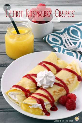 Side view of two lemon and raspberry crepes garnished with whipped cream on a white plate with a glass of orange juice in the background. 