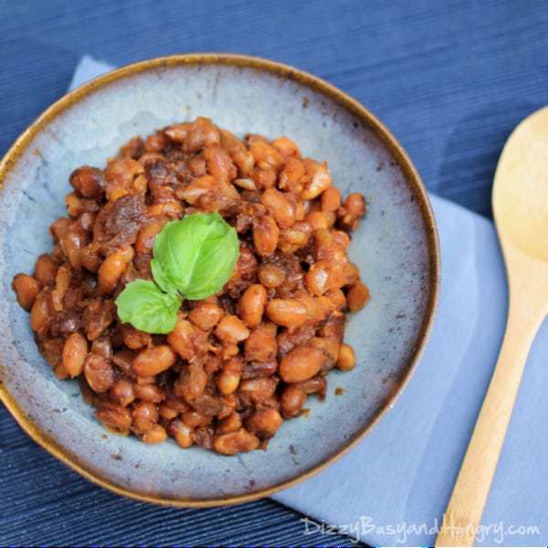 Overhead shot of Boston baked beans garnished with herbs in a rustic blue bowl and a blue background with a wooden spoon. 