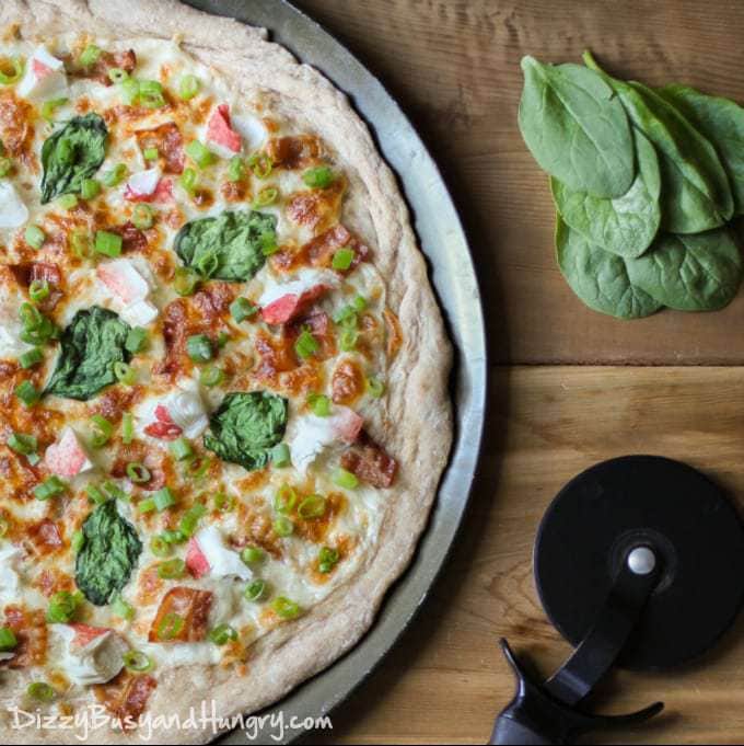 Overhead shot of crab bacon spinach pizza on a black plate on a wooden table with spinach and a pizza cutter in the background. 