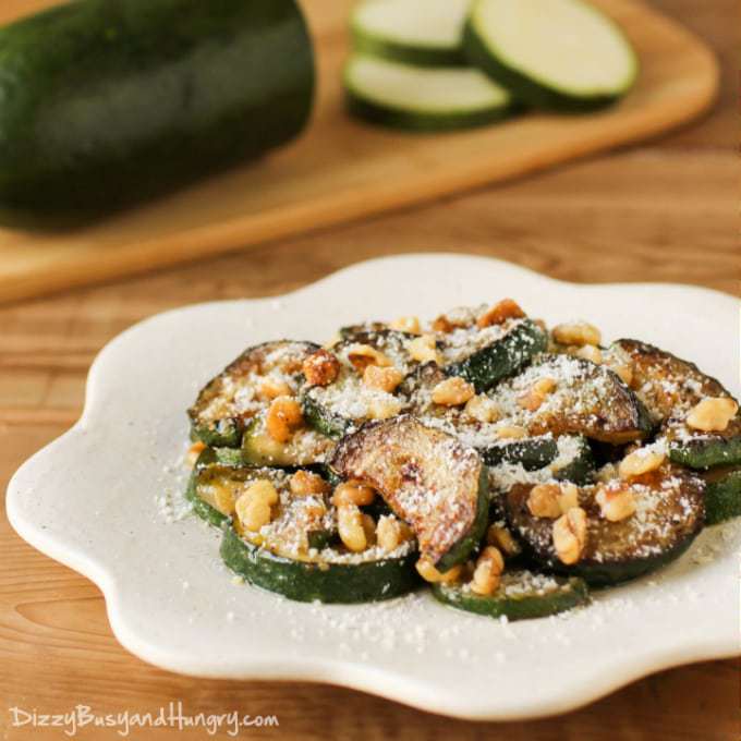 Side view of sautéed zucchini with walnuts on a white plate and a half sliced zucchini in the background on cutting board.