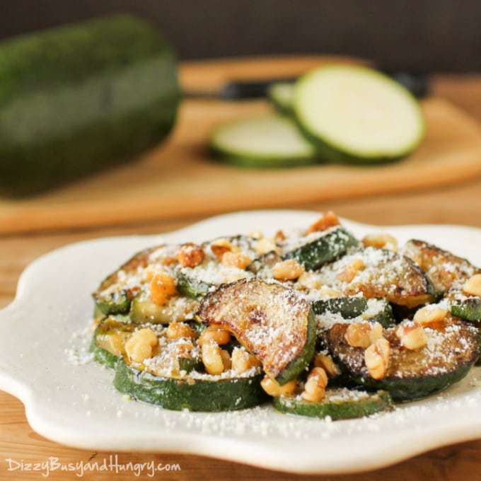 Front view of sautéed zucchini on a white plate with a half sliced zucchini in the background on a cutting board. 