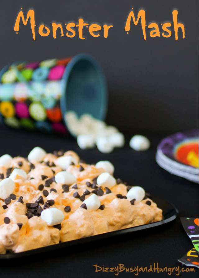 Close up shot of monster mash in a black pan with mini marshmallows and halloween decorations in the background. 