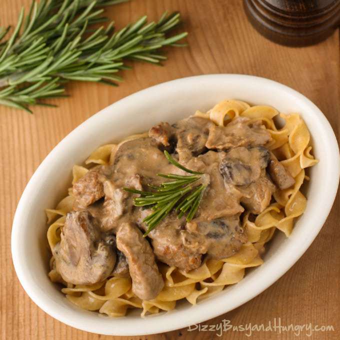 Overhead shot of crockpot beef stroganoff in a white oval bowl on a wooden table with herbs in the background. 