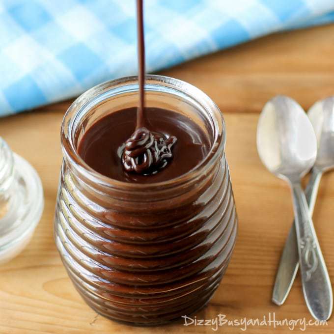 Side view of chocolate sauce being poured into a clear jar with a blue and white plaid cloth and spoons in the background.