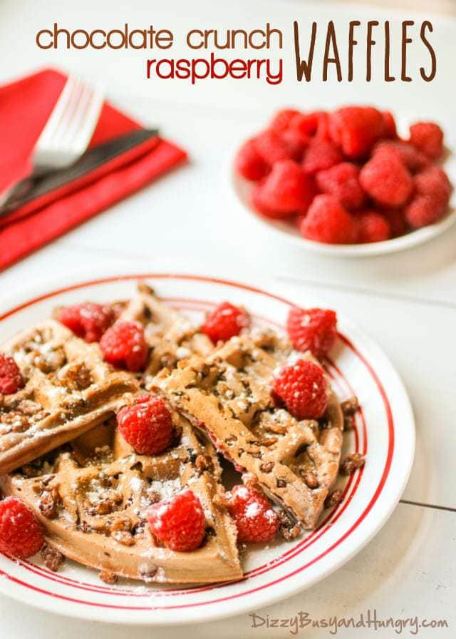 Side view of chocolate crunch raspberry waffles sprinkled with powdered sugar and raspberries on a white plate with a bowl of raspberries in the background. 