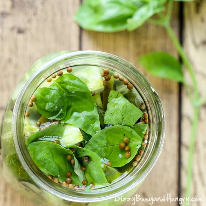 Overhead shot of jalapeño garlic basil pickles in a clear mason jar on a wooden surface with herbs on the side.