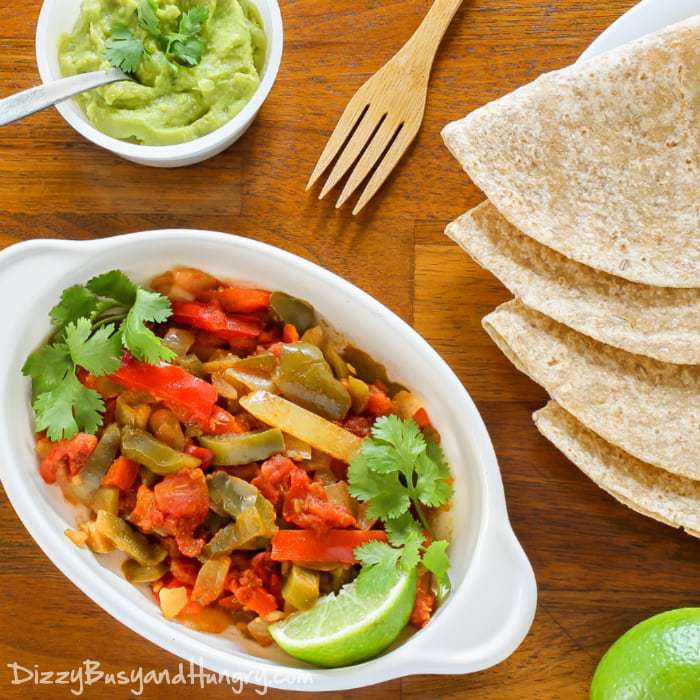 Overhead shot of veggie fajita mix in a white bowl with layered tortillas and a bowl of guacamole in the background. 