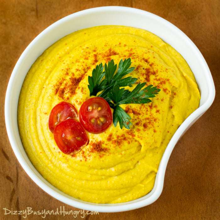 Overhead shot of turmeric hummus in a white bowl garnished with tomatoes and herbs. 