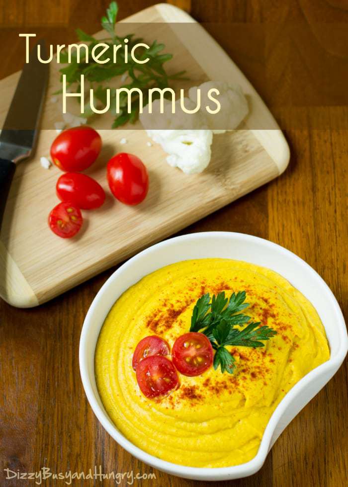 Turmeric hummus sprinkled with herbs and sliced tomatoes with a wooden cutting board with ingredients in the background. 