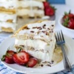 Side shot of a piece of maple bacon waffle ice cream cake on a white plate with a fork and sliced strawberries on the side.