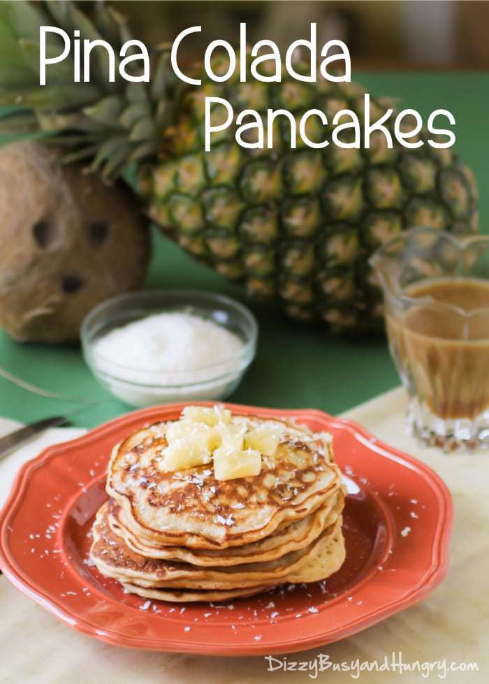 Side shot of stacked tropical pancakes garnished with pineapple and shaved coconut on a red plate. 