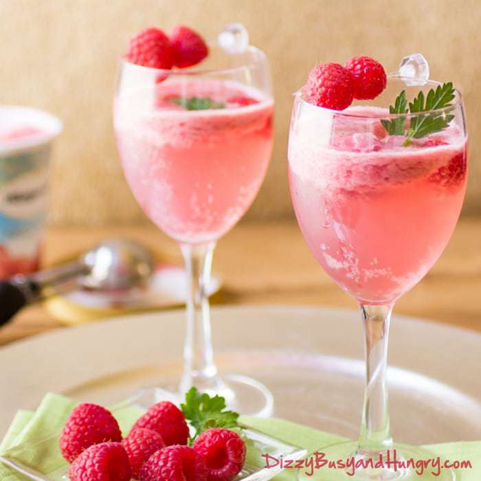 Side shot of raspberry sorbet wine spritzer in two wine glasses garnished with herbs and raspberries on a green cloth. 