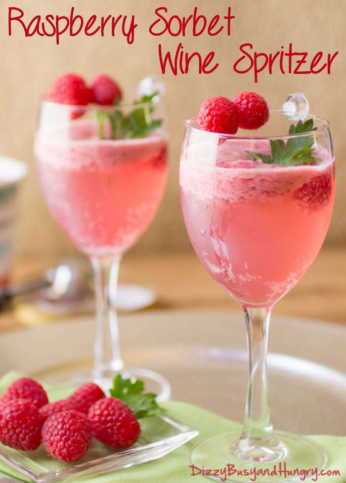 Side shot of raspberry sorbet wine spritzer in two wine glasses garnished with herbs and raspberries on a green cloth. 