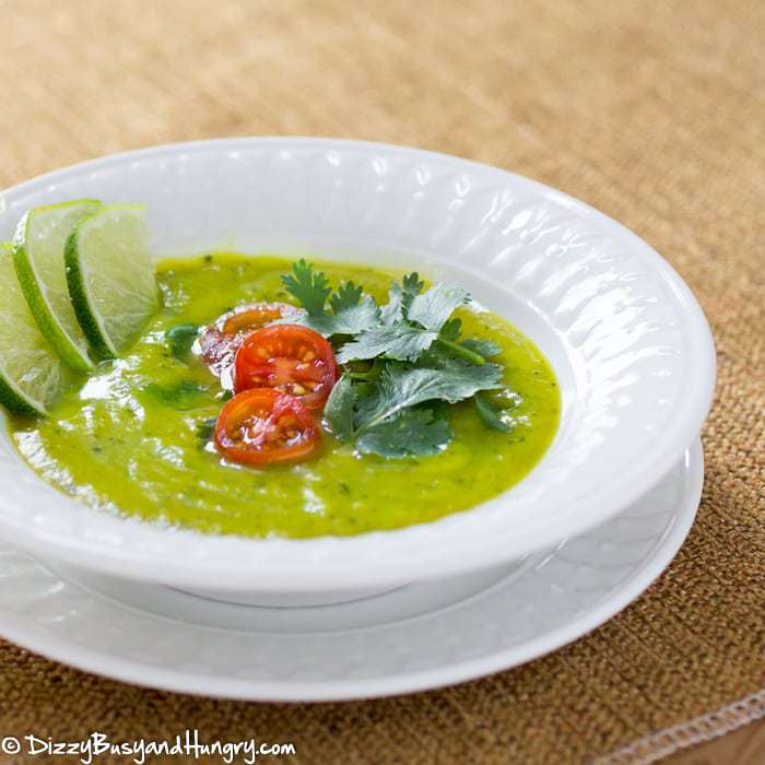 Side view of zucchini curry lime soup garnished with herbs tomatoes and sliced lime in a white bowl on a white plate. 