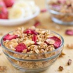 Close up shot of strawberry granola in a clear owl with sliced bananas and strawberries in the background.