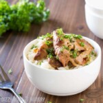 Side view of crock pot chicken thighs in a white bowl on a wooden surface with a fork and herbs in the background. 