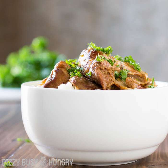 Side shot of crock pot chicken thighs in a white bowl on a wooden surface with herbs and a fork in the background. 