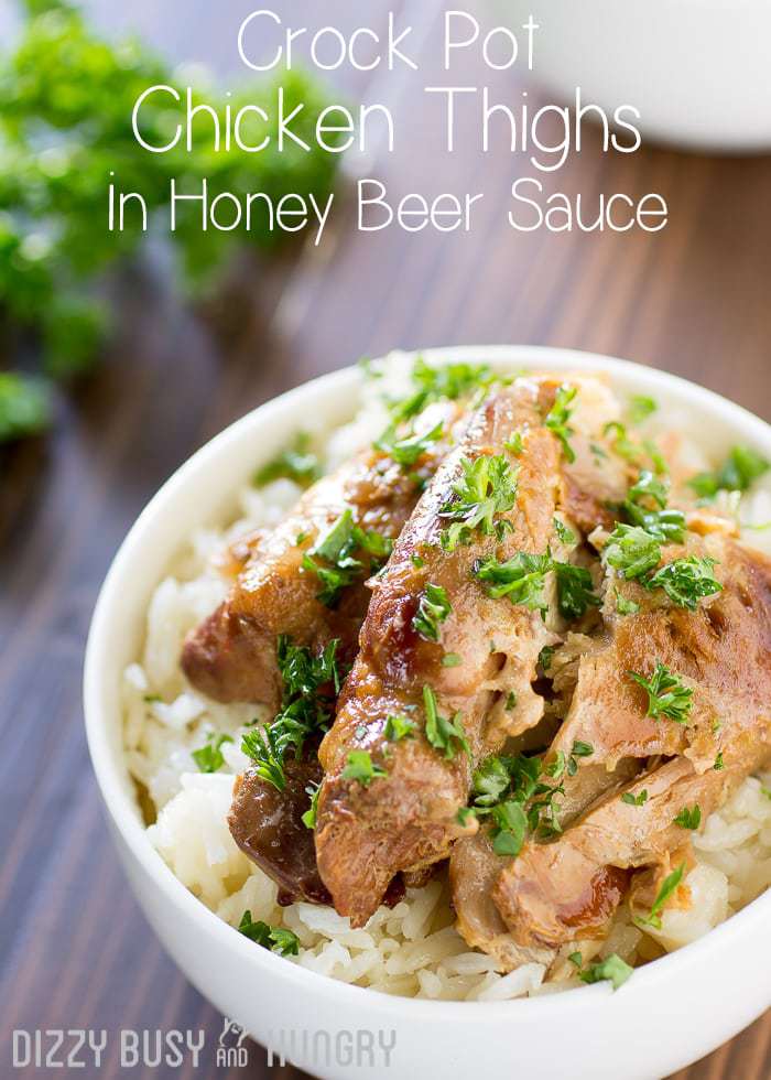 Close up view of crock pot chicken thighs in a white bowl on a wooden surface with herbs and a fork in the background. 