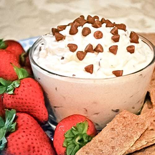 Close up shot of cinnamon cannoli dip in a clear bowl surrounded by graham crackers and strawberries on a blue plate. 