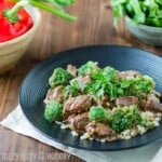 Side view of crock pot beef and broccoli on a black plate on a white cloth with peppers and herbs in the background.