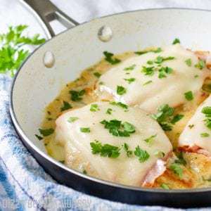 Side view of chicken cordon bleu in a white and black pan on a blue plaid cloth sprinkled with herbs.