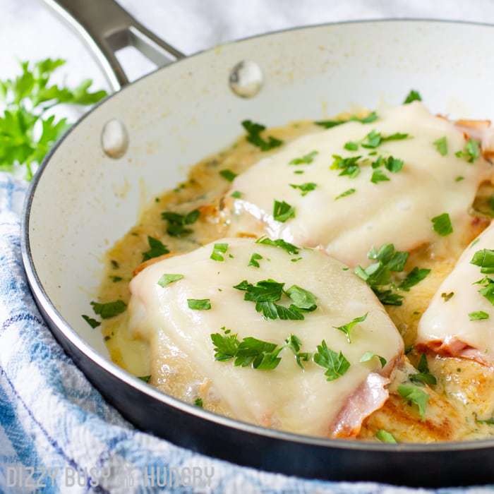 Side view of chicken cordon bleu in a white and black pan on a blue plaid cloth sprinkled with herbs. 