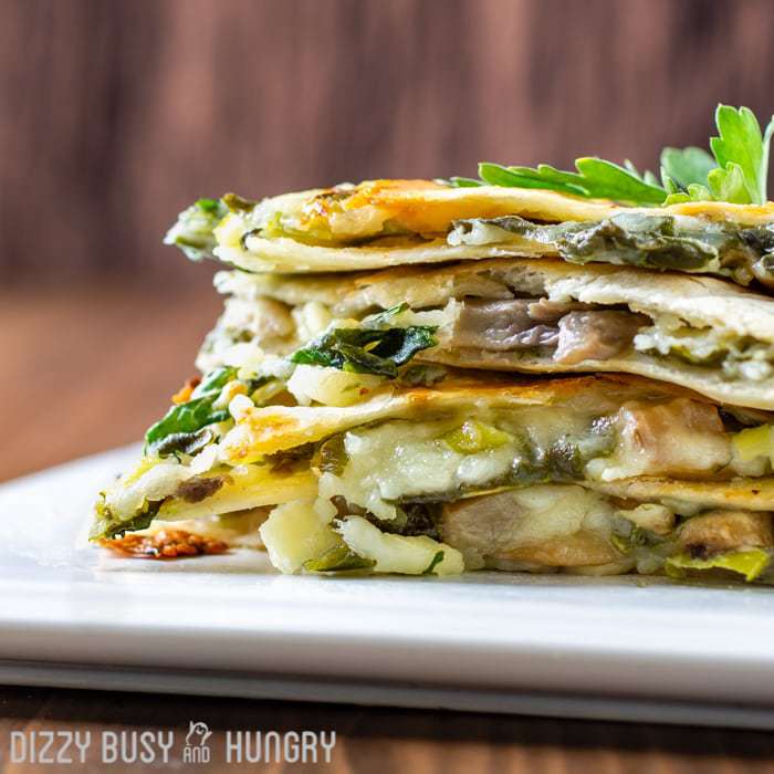 Side view of multiple spinach mushroom quesadillas stacked on a white plate with herbs in the background. 