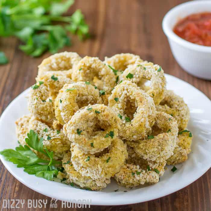 Side view of baked calamari with herbs on a white plate with a bowl of sauce in the background.