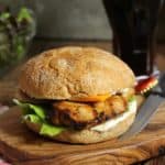 Front view of a grilled chicken burger on a wooden cutting board with a knife on the side.