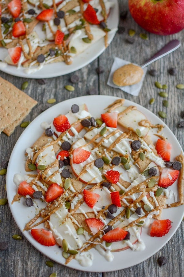 Top view of apples, strawberries, graham crackers, pepitas, chocolate chips on a white plate - after school snacks