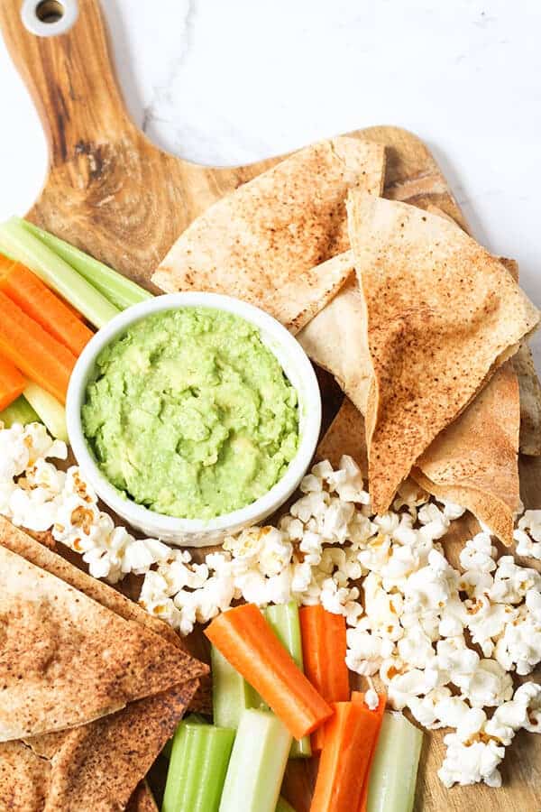 Top view of pita chips surrounded by veggies, popcorn and a green dip