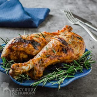 Side view of two turkey legs garnished with rosemary on a small blue plate with a blue napkin, fork and knife behind.
