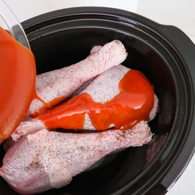 Close up shot of sauce being poured over raw, seasoned turkey legs in black crock pot.