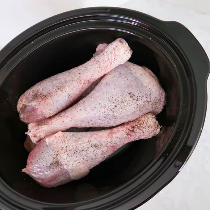 Process shot of raw, seasoned turkey legs in black crockpot.