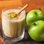 Smoothie with cinnamon, cut up apples, and a cinnamon stick on top, in a clear glass, beside two granny smith apples.