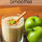 Close up shot of smoothie with cinnamon, cut up apples, and a cinnamon stick on top, in a clear glass, beside two granny smith apples.