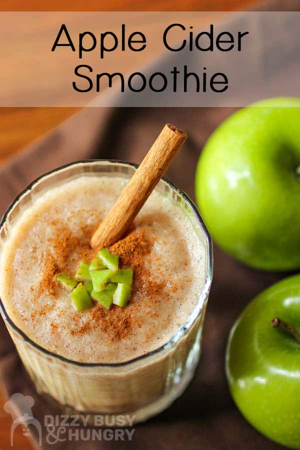 Close up shot of smoothie with cinnamon and a cinnamon stick on top, in a clear glass, beside two granny smith apples.
