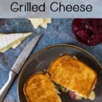Overhead view of sliced halves of brie grilled cheese with a knife, sliced cheese, and cranberry sauce on a blue background and plate.