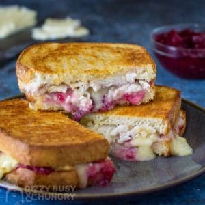 Side view of three stacked halves of brie grilled cheese with cranberry sauce and sliced cheese on a blue background.