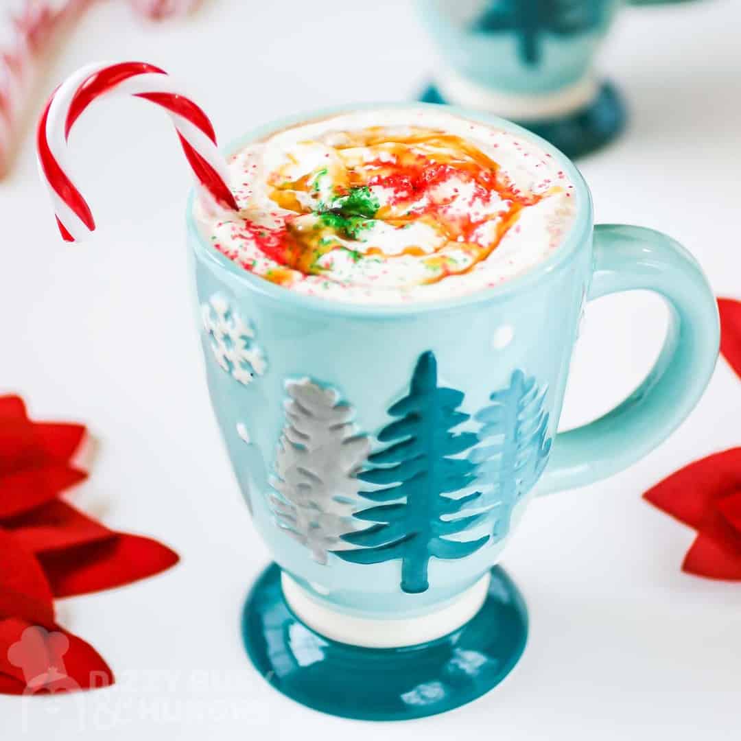 Close up shot of hot chocolate with whipped cream and a candy cane in a blue mug with a white background.
