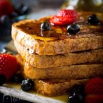 Close up shot of four stacked french toast covered in syrup, and garnished with strawberries and blueberries.