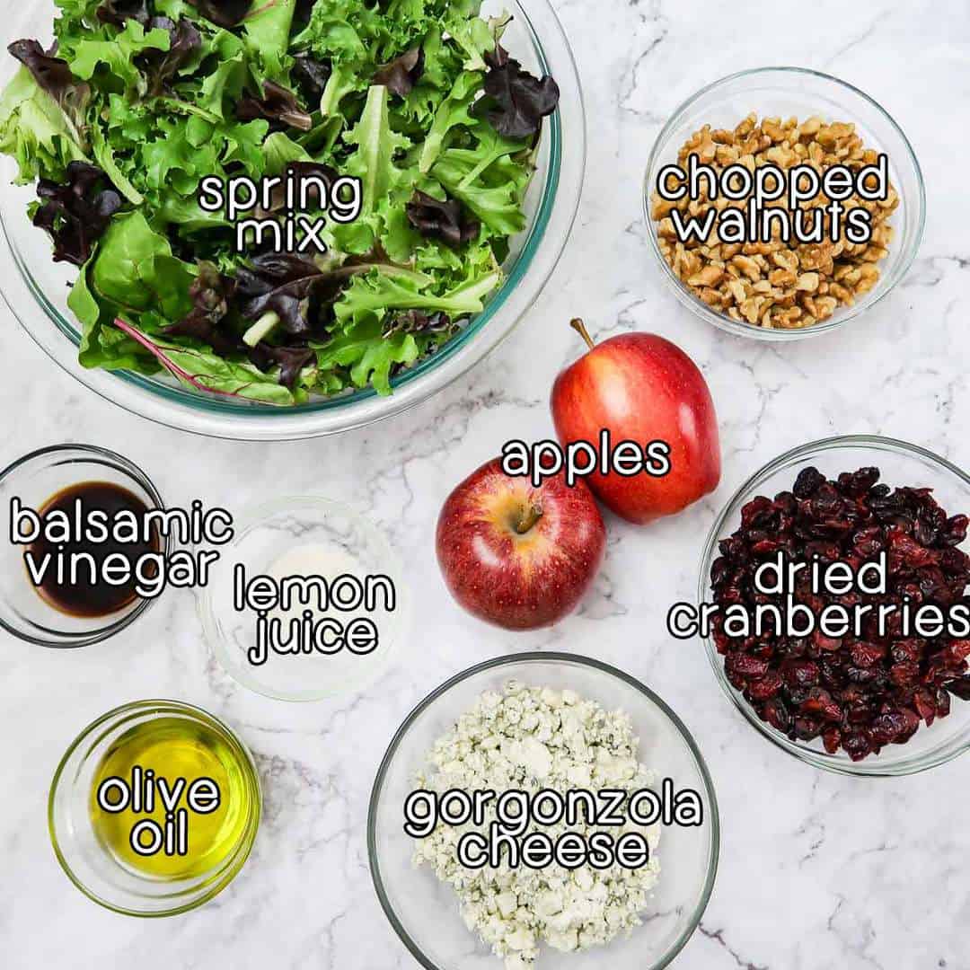 Overhead view of ingredients- spring mix, chopped walnuts, apples, dried cranberries, gorgonzola cheese, lemon juice, olive oil, and balsamic vinegar.