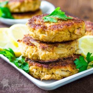 Side view of three crab cakes stacked on a white plate garnished with herbs and sliced lemons.