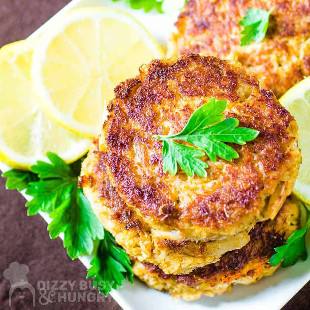 Overhead shot of three crab cakes stacked on a white plate garnished with herbs and sliced lemons.