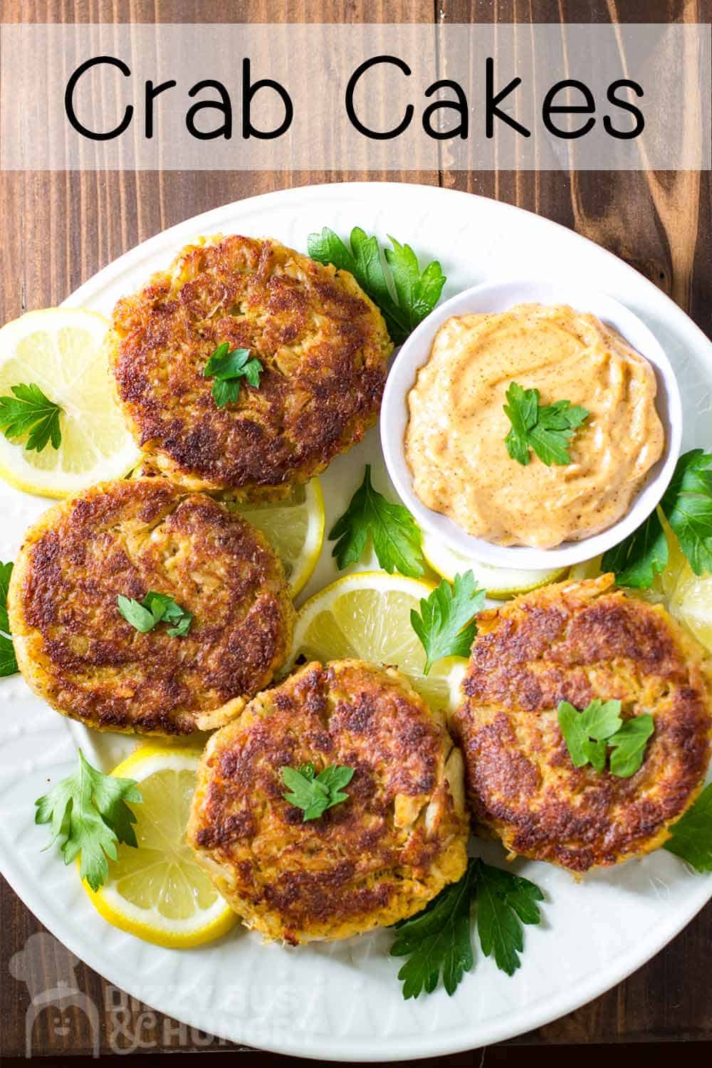 Overhead shot of four crab cakes in a circle on a white plate with a bowl or dipping sauce on the side on a bed of sliced lemons and herbs.