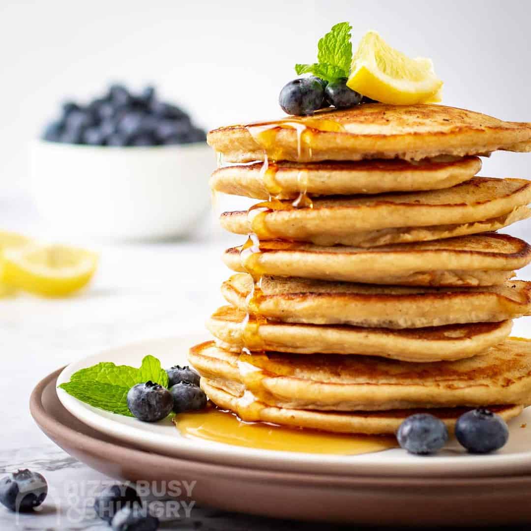 Side view of multiple lemon pancakes stacked on a white plate garnished with mint, lime, syrup, and blueberries.